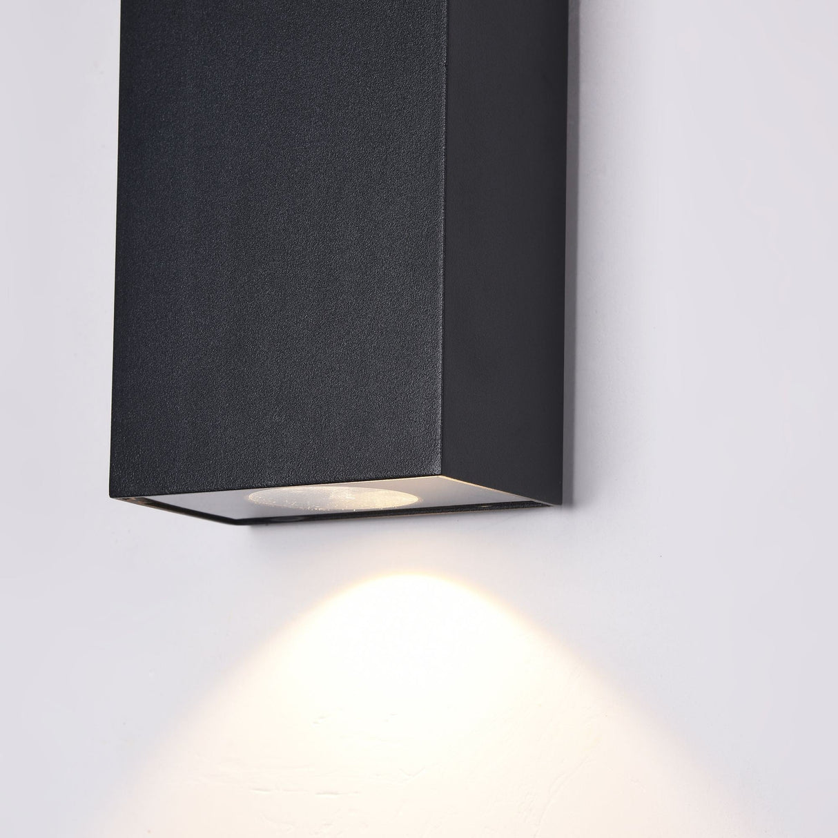 The Times Square Outdoor Wall Light 3000K 6W in black is a wall-mounted rectangular fixture with modern LED lighting, casting a warm glow on white walls. Its sleek design and downward-facing bulb bring contemporary elegance to any space.
