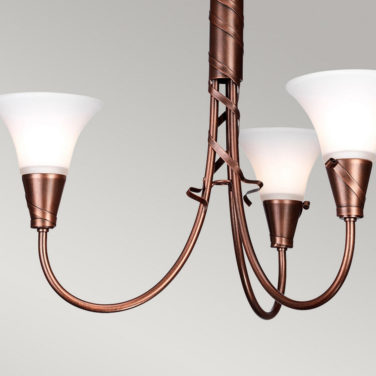 The Emily 3 Light Chandelier - Copper Patina exudes vintage charm with its traditional bronze design and three white, cone-shaped glass shades. Its elegantly curved metal arms feature decorative spirals and a striking copper patina finish, making it stand out beautifully against a neutral gray background, ideal for vintage interiors.