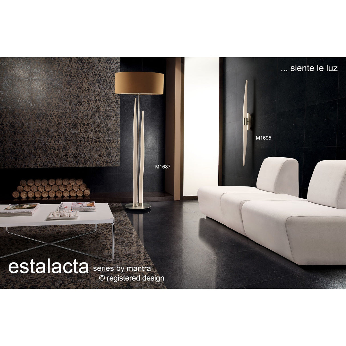 Modern living room featuring the Estalacta 3 Light Floor Lamp - Silver with Black Shade, a white sectional sofa, white coffee table, wall sconce, stacked firewood, and textured dark wall. Text reads "Estalacta series by Mantra… siente la luz.