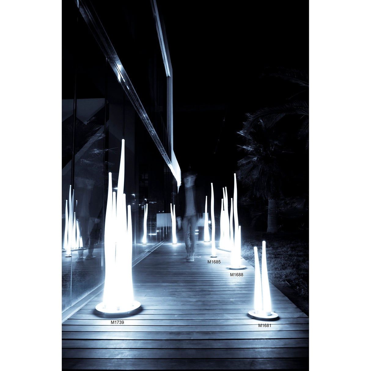 A person walks along a wooden path at night, illuminated by Estalacta 1 Light Small Floor Lamps in silver and opal white. The sleek, modern building on the left reflects their glow, with trees visible in the dark background.
