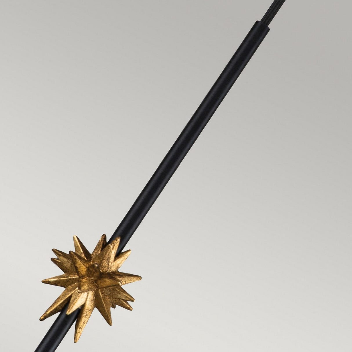 A metallic starburst adorns the lower part of a matte black pole, reminiscent of the Etoile 1 Light Medium Pendant. Set against a light gray background, this starburst showcases sharp, radiating points with a lustrous golden finish that strikingly contrasts with the pole's simple design.