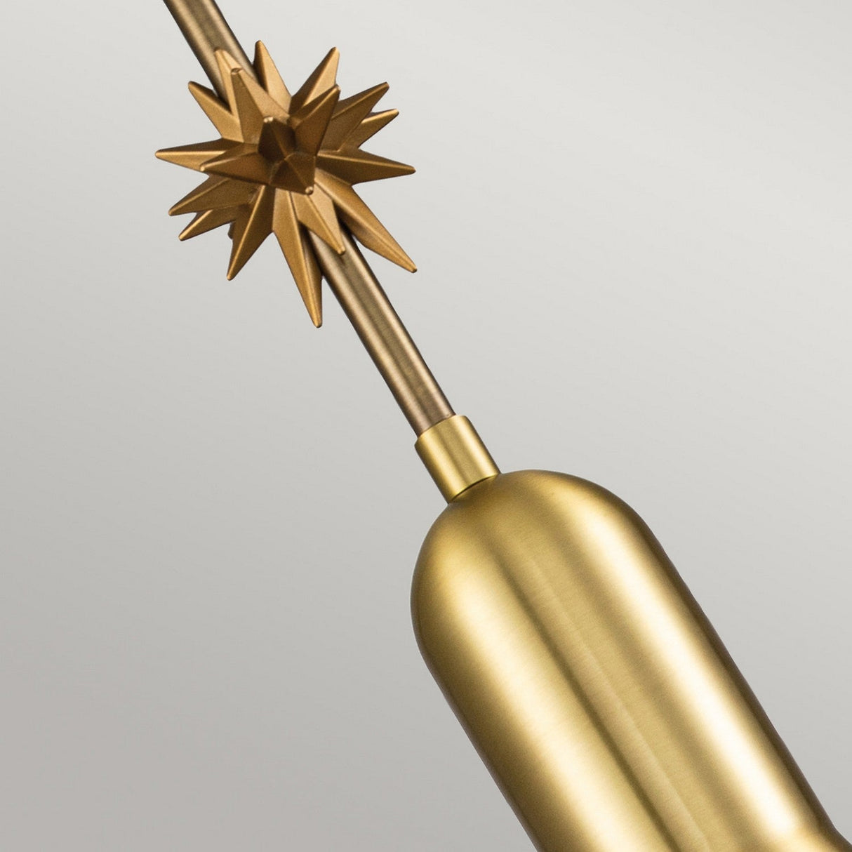 A close-up view of the Etoile 1 Light Small Pendant Light in aged brass, featuring a cylindrical base and an angular star-shaped ornament on the stem against a plain gray background. The elegant design exudes vintage charm, showcasing its refined craftsmanship.