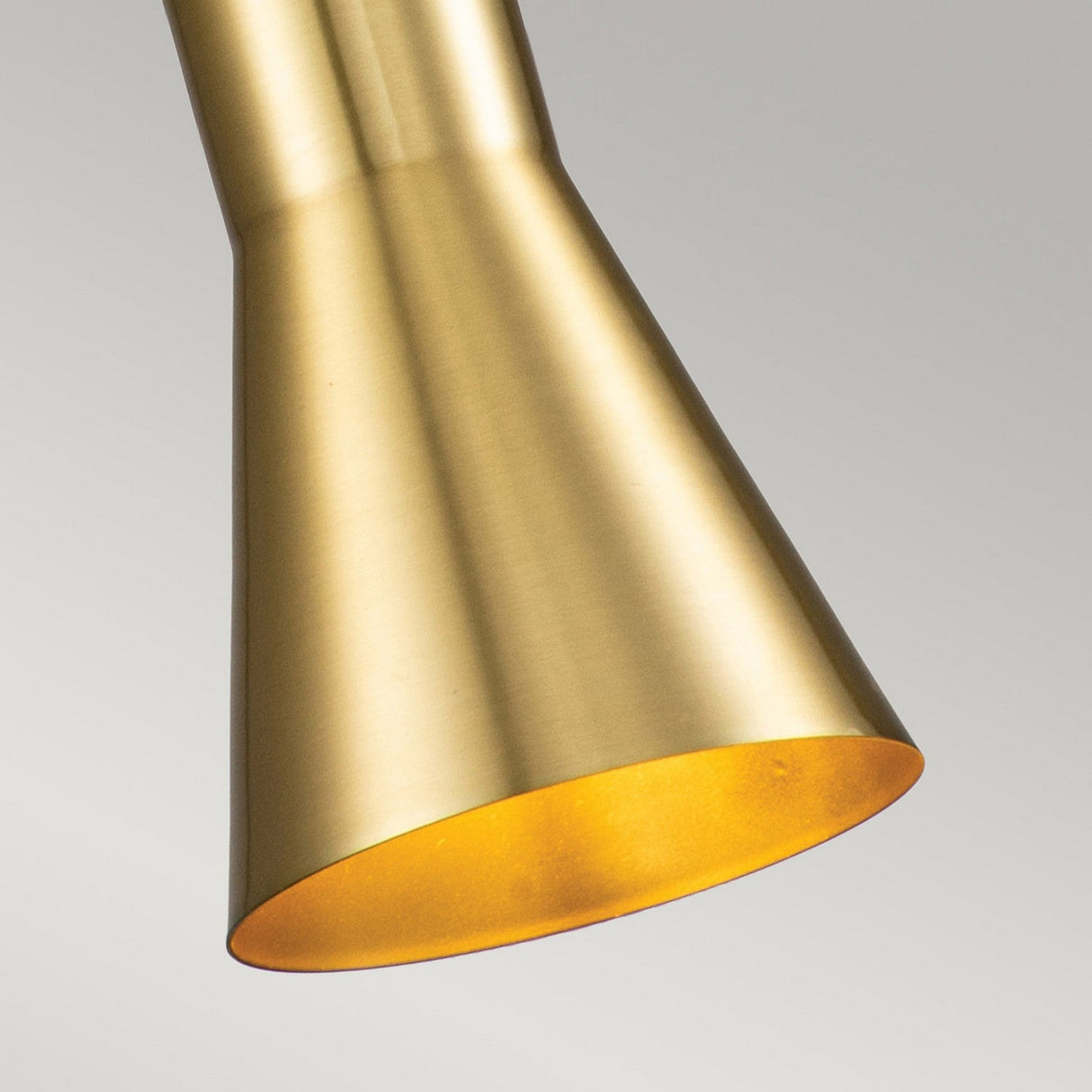 A close-up shot of the Etoile 1 Light Small Pendant Light in Aged Brass reveals its smooth, matte finish. It hangs from an adjustable black cord against a simple backdrop, with its cone-shaped fixture angled downwards to highlight its sleek design and metallic sheen.