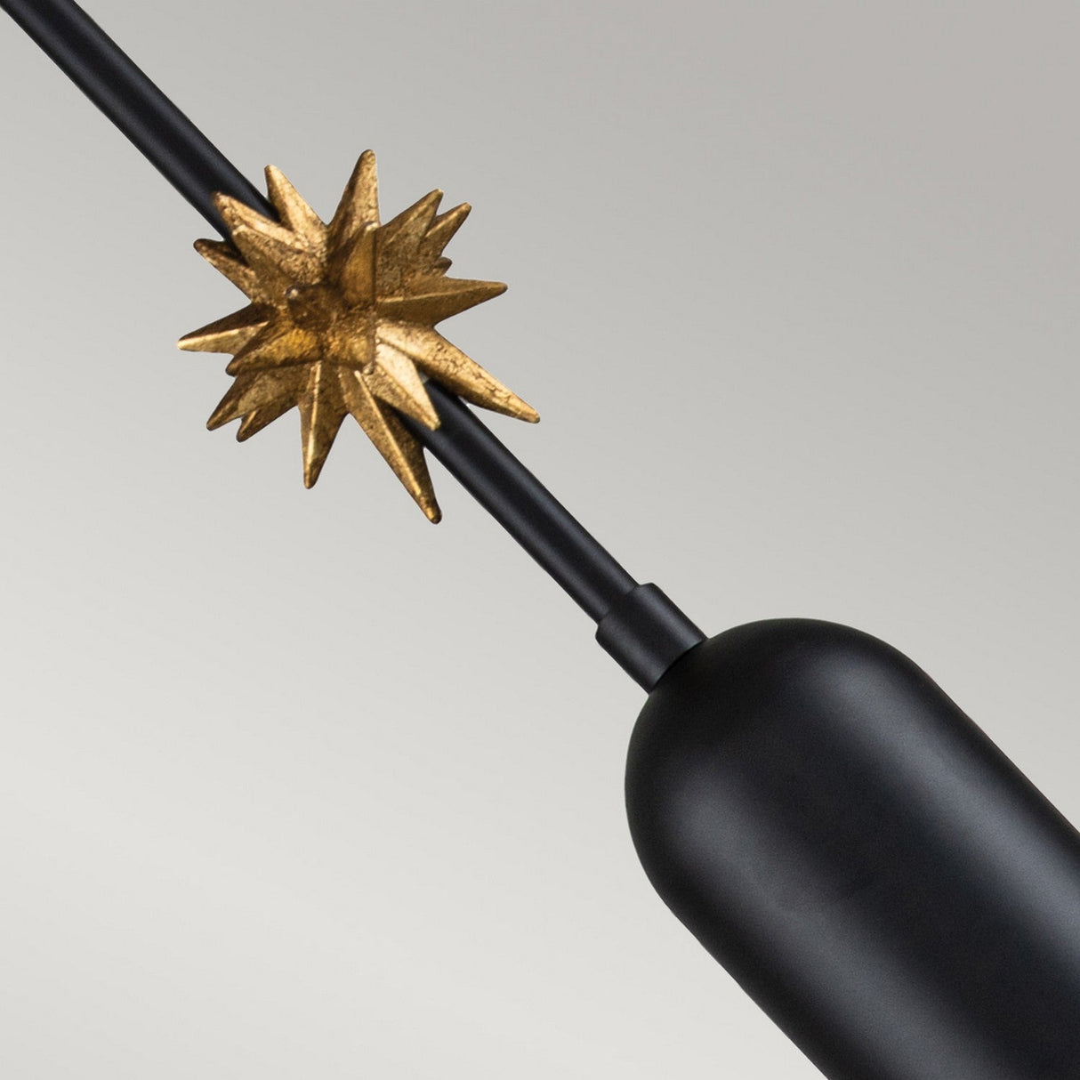The Etoile 1 Light Small Pendant - Matte Black showcases a sleek, cylindrical design featuring a long, thin rod extending diagonally. A small gold starburst adorns the rod, adding a touch of elegance to this modern lighting fixture against the neutral gray background.