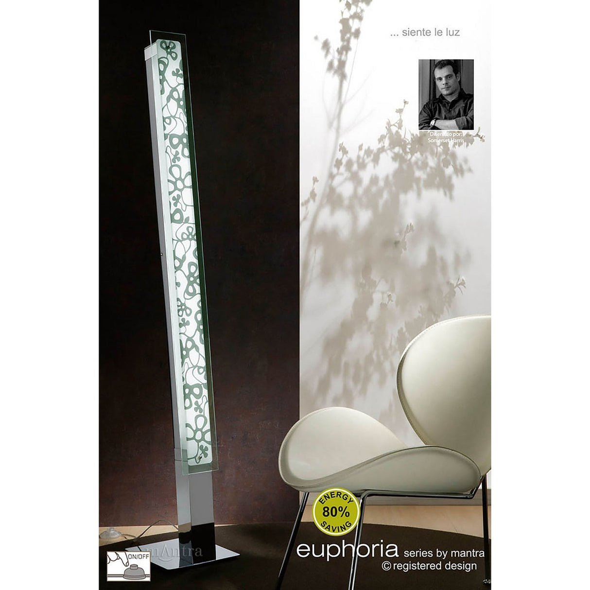 The Euphoria 2 Light Floor Lamp - Chrome, featuring a tall contemporary design with a decorative floral pattern, stands beside a uniquely shaped white chair against a shadowed wall. A black-and-white photo and Spanish text appear in the upper right corner.