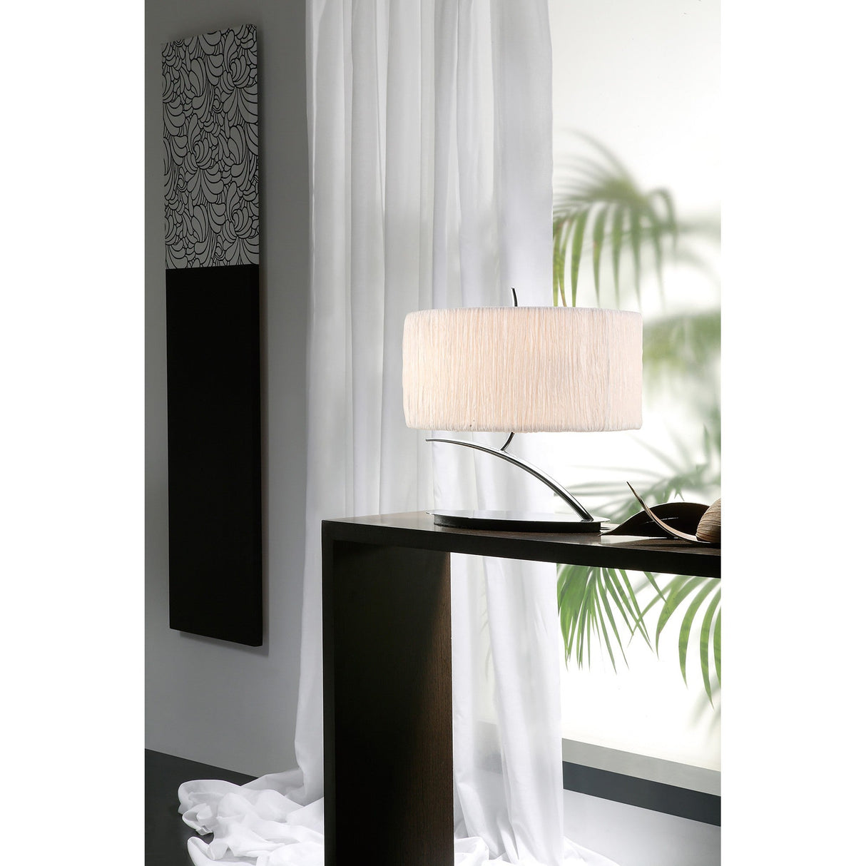 A modern room features sheer white curtains, a black console table, and the Eve 2 Light Table Lamp in polished chrome with a white oval shade. Leafy plant shadows fall on the wall beneath striking black-and-white wall art.