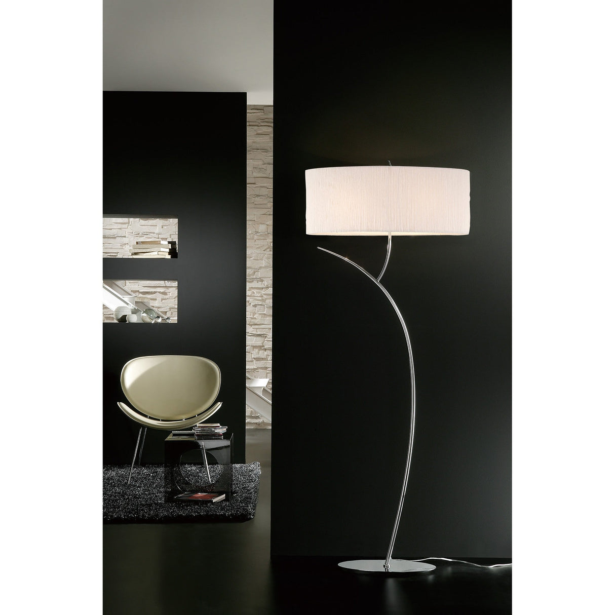 The Eve 2 Light Floor Lamp in chrome with a white oval shade stands on a dark floor beside a green chair and small table in a modern room featuring dark and stone-textured walls.