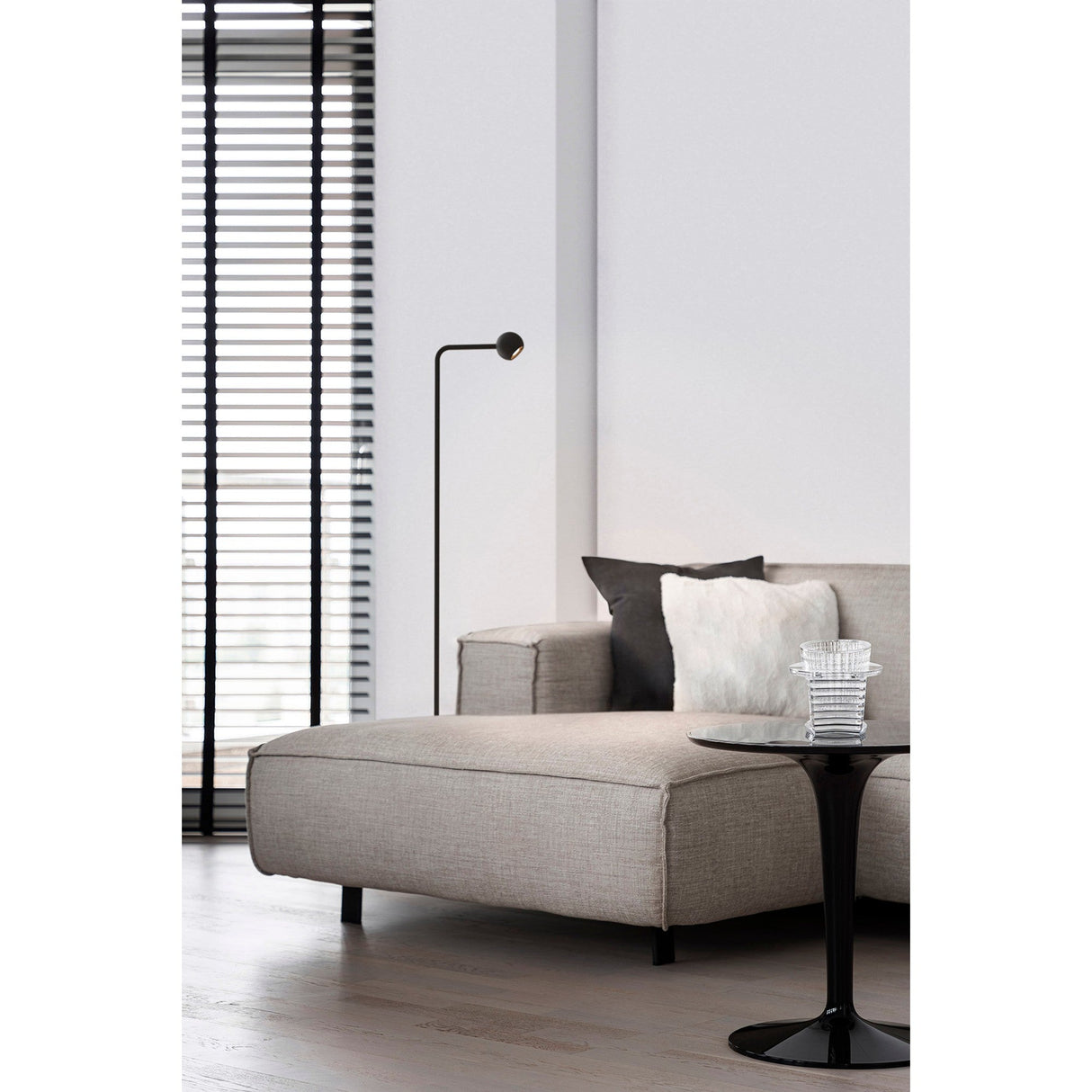 A modern living room featuring a light gray sectional sofa, black and white cushions, a black side table with a glass vase, the Eyes LED Floor Lamp 6W 3000K - Sand Black, and large windows with black blinds letting in natural light.