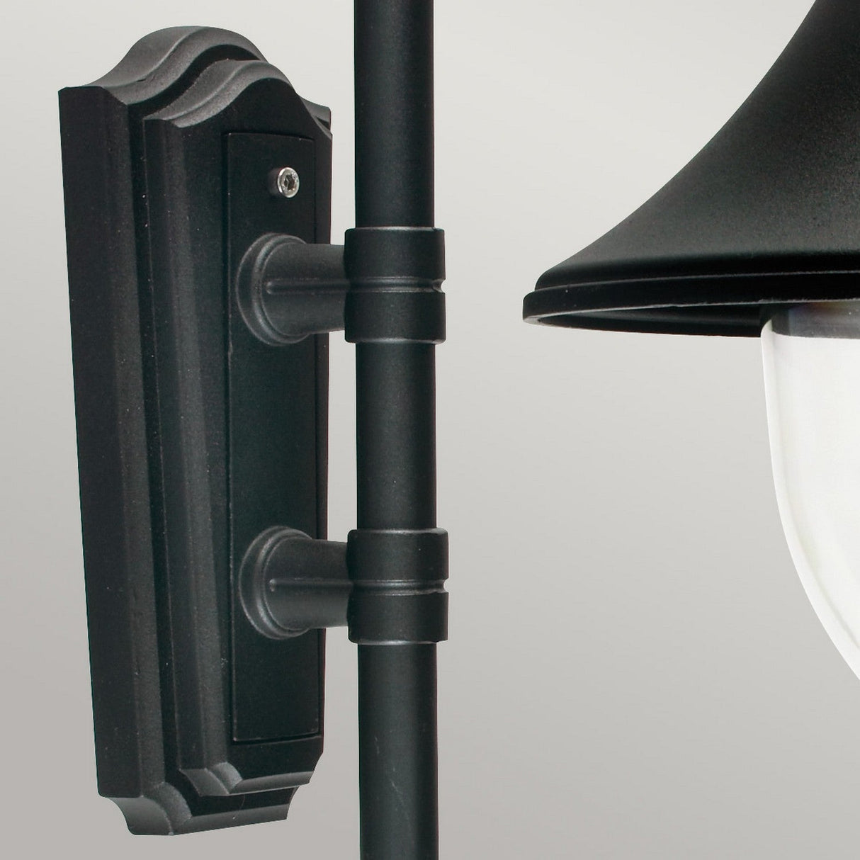 A close-up view of a black outdoor wall lantern showcases a classic design. The fixture, reminiscent of the Firenze Outdoor 1 Light Wall Lantern - Black, features a decorative backplate and pole extending to a hooded lamp with frosted glass shade, set against a neutral background.