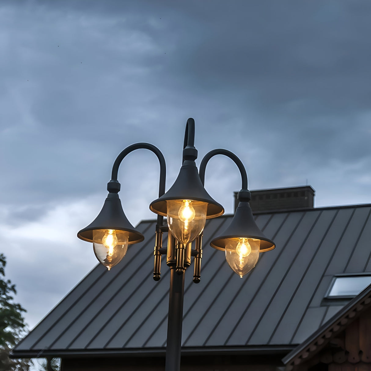 The Firenze 3 Light Lamp Post - Black, featuring a sleek black finish, casts its illumination against a moody, cloudy sky. This elegant three-headed street lamp stands tall as a building with a sloped metal roof is partially visible in the background.