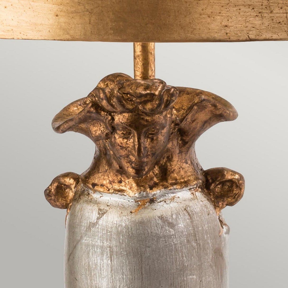 The Bienville 1 Light Table Lamp - Silver/Gold and Brown features a sophisticated sculpted base with an ornate design, including a face and decorative elements. Its modern aesthetic is highlighted by a textured metallic finish that combines rich gold tones with subtle silver accents for an elegant appearance.