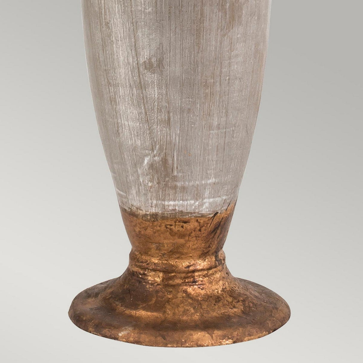 A close-up of the Bienville 1 Light Table Lamp showcases its metallic vase base with a textured, rough surface. It features a bronze-colored bottom that gradually transitions to a lighter silver tone towards the top, highlighting its modern style. The background is a soft gradient gray.