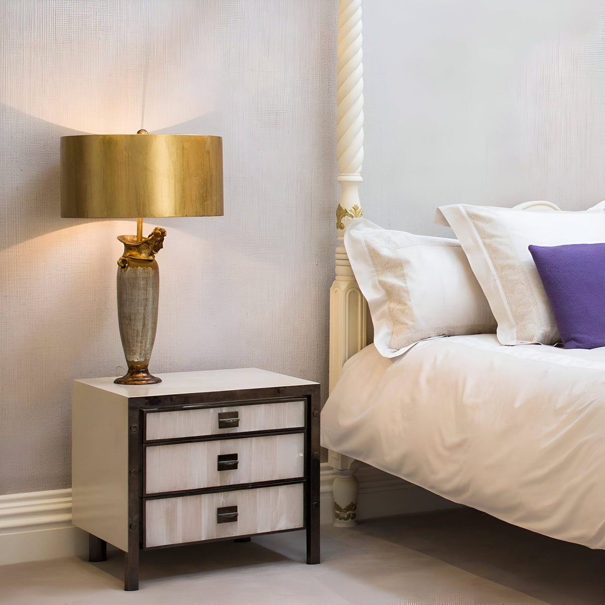 A cozy bedroom scene showcases a white bed, a purple pillow, and beige sheets. The Bienville 1 Light Table Lamp - Silver/Gold and Brown rests on a three-drawer bedside table, casting a modern glow on the textured gray wall.