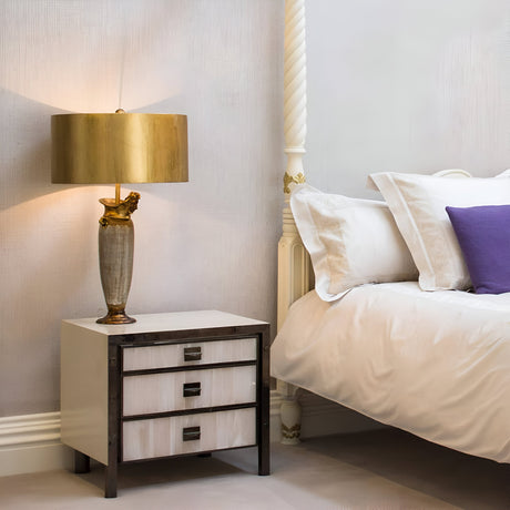 A cozy bedroom scene showcases a white bed, a purple pillow, and beige sheets. The Bienville 1 Light Table Lamp - Silver/Gold and Brown rests on a three-drawer bedside table, casting a modern glow on the textured gray wall.