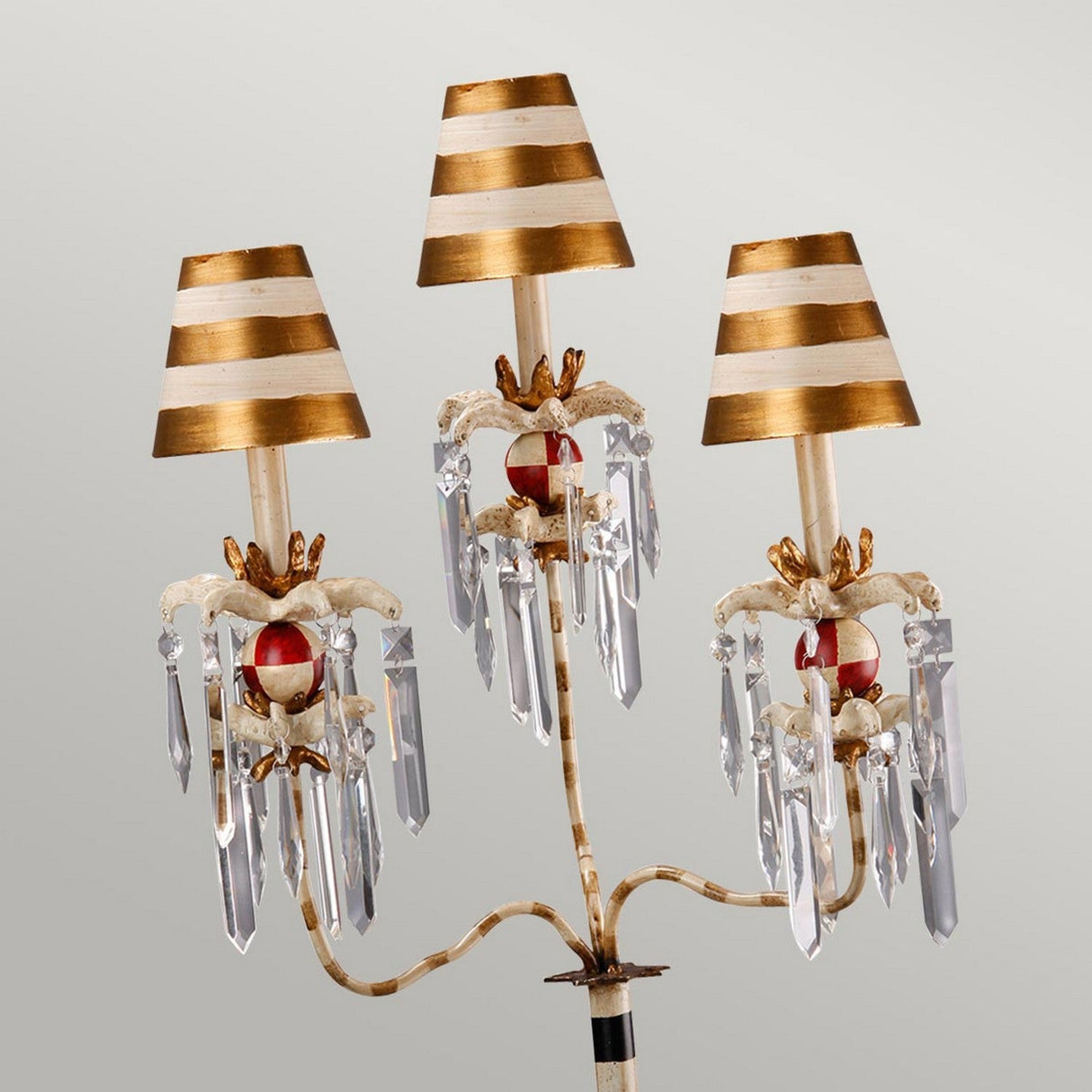 The Birdland 3 Arm Table Lamp - Cream & Gold features a classic candelabra-style design with three candle-like arms. Each arm is adorned with a striped gold and white lampshade, accented by red details and clusters of K9 cut glass, creating an elegant and timeless look.