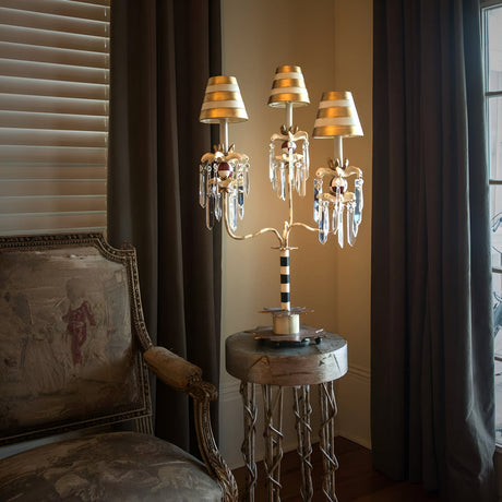 A luxurious room featuring an ornate chair with upholstered fabric is complemented by a round side table. On the table sits a Birdland 3 Arm Table Lamp in Cream & Gold, adorned with candelabra-style arms and K9 cut glass clusters. Two large windows with heavy curtains let soft lighting flow into the space.