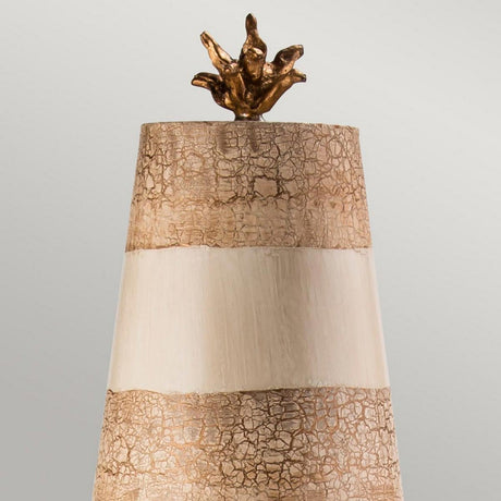 The Birdland 1 Light Table Lamp in cream and gold features two beige, textured bands and a smooth central band adorned with K9 cut glass accents. It is elegantly topped with a small, abstract golden sculpture that resembles a star or flower, set against a plain gray backdrop.