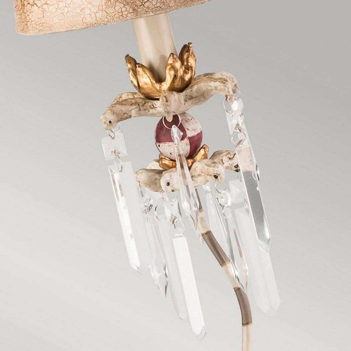 The Birdland 1 Light Table Lamp - Cream & Gold features a decorative beige lampshade accented with gold and a maroon orb, complemented by clear K9 cut glass accents that dangle elegantly. Its ornate appearance is further enhanced by a crackled putty finish backdrop.