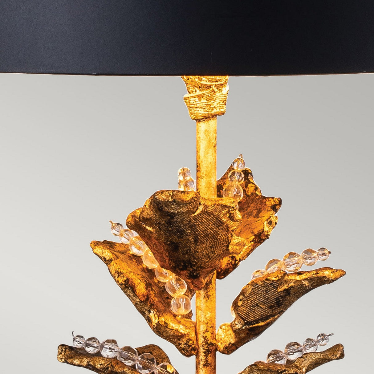 The Camilia 1 Light Table Lamp in Antique Gold & Black features a black shade paired with a gold base designed to resemble plant leaves. The elegant gold leaf design is accentuated by stems embellished with clear crystal beads, offering a sophisticated and decorative touch to any room.