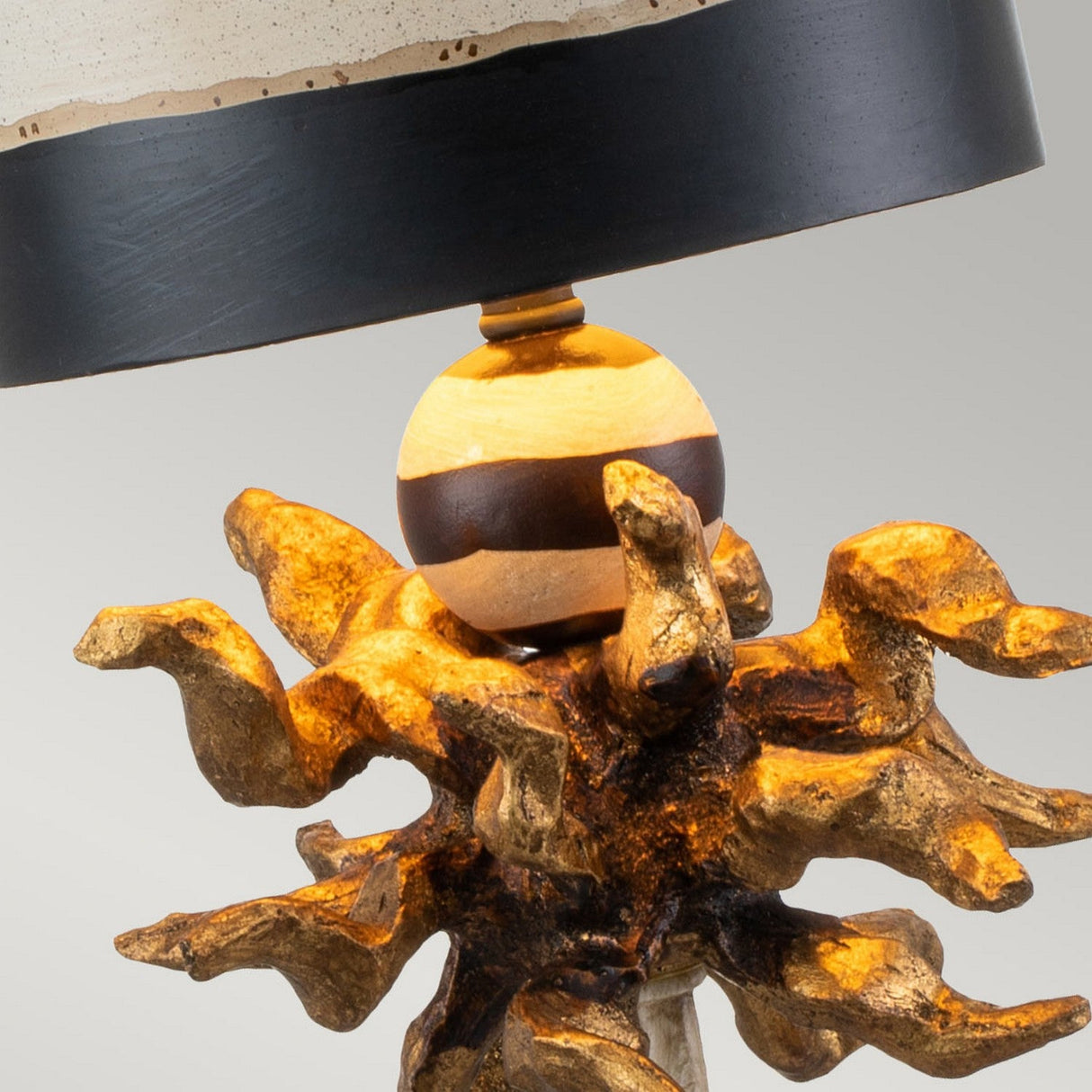 The Dominique 1 Light Table Lamp boasts a contemporary design with a gold leaf base that mimics organic, twisted roots. It includes a round wooden sphere that supports a black drum shade accentuated by cream stripes, all presented against a neutral background.