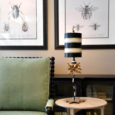 A cozy room with a green upholstered chair next to a round wooden table featuring the Dominique 1 Light Table Lamp in gold leaf, black, and cream stripes. Framed vintage insect illustrations on the wall add a classic touch to the decor, while books are neatly arranged on a lower shelf.