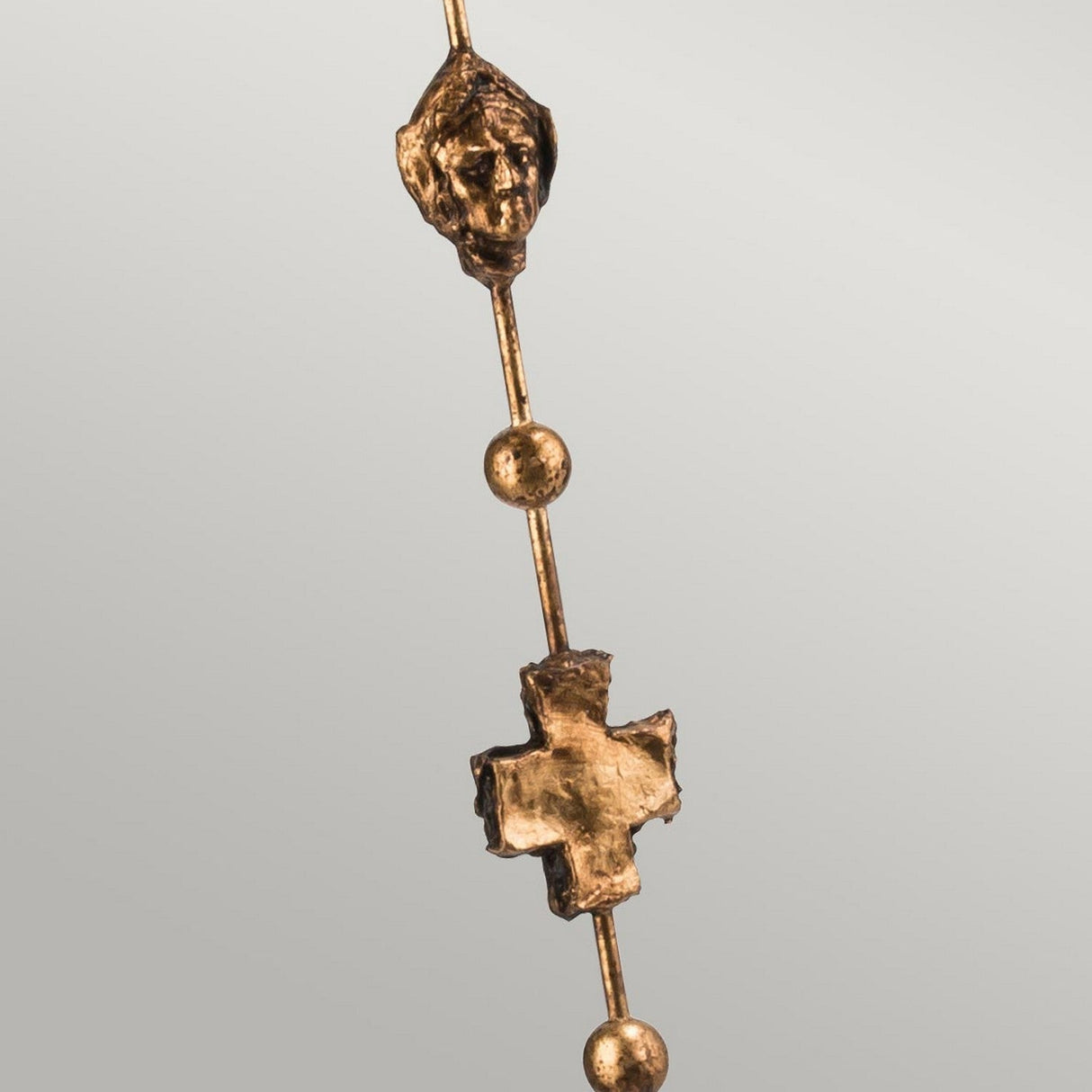 The Fragment 1 Light Floor Lamp in an aged gold finish features a pendant with a stylized human head, two spherical beads, and a cross-shaped element arranged vertically. The intricate design is elegantly highlighted against the plain background as if illuminated by ambient lighting.
