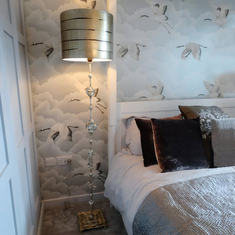 A bedroom showcases the Fragment 1 Light Floor Lamp in an aged gold finish, featuring an ornate design and decorative stem, positioned beside a bed adorned with textured pillows. The wallpaper displays flying cranes set against a cloudy sky, while a wardrobe panel peeks from the left, contributing to the room's ambient lighting.