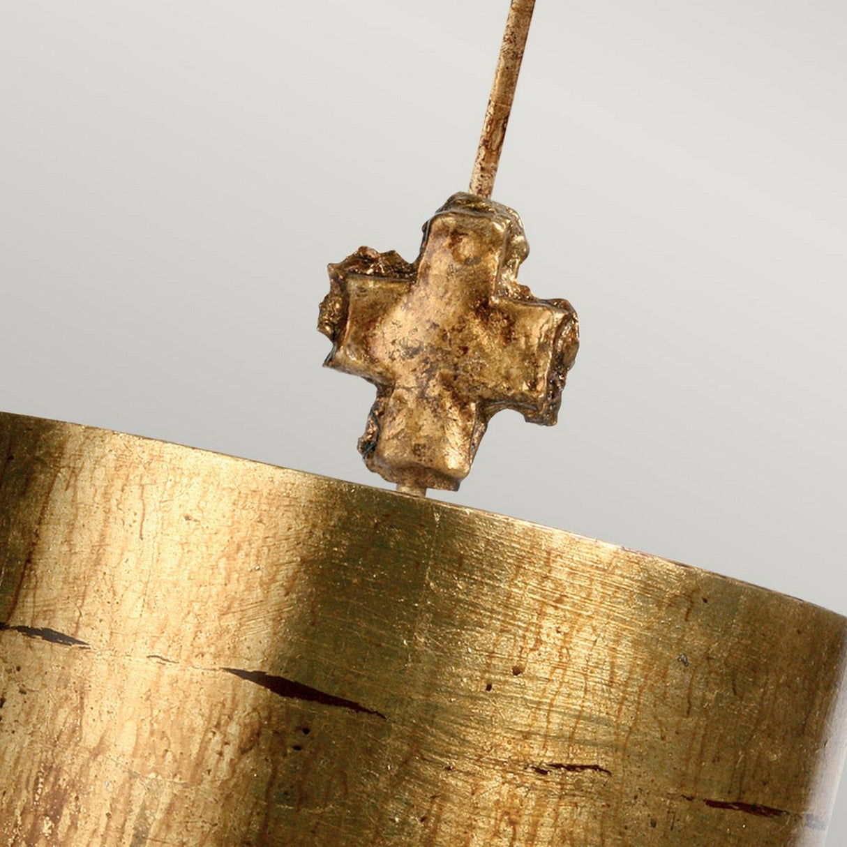 A detailed view of the Fragment 1 Light Pendant in aged gold displays its small, rustic cross design on a thin wire. In the background is a large cylindrical piece with a textured surface, enhancing the metallic and warm ambiance.
