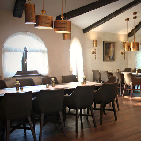 A cozy dining room with rustic wooden beams and soft natural lighting features elegant tables set with green chairs and small floral centerpieces. The decor includes draped windows, a Fragment 1 Light Pendant with an aged gold finish, and a framed artwork on the wall.