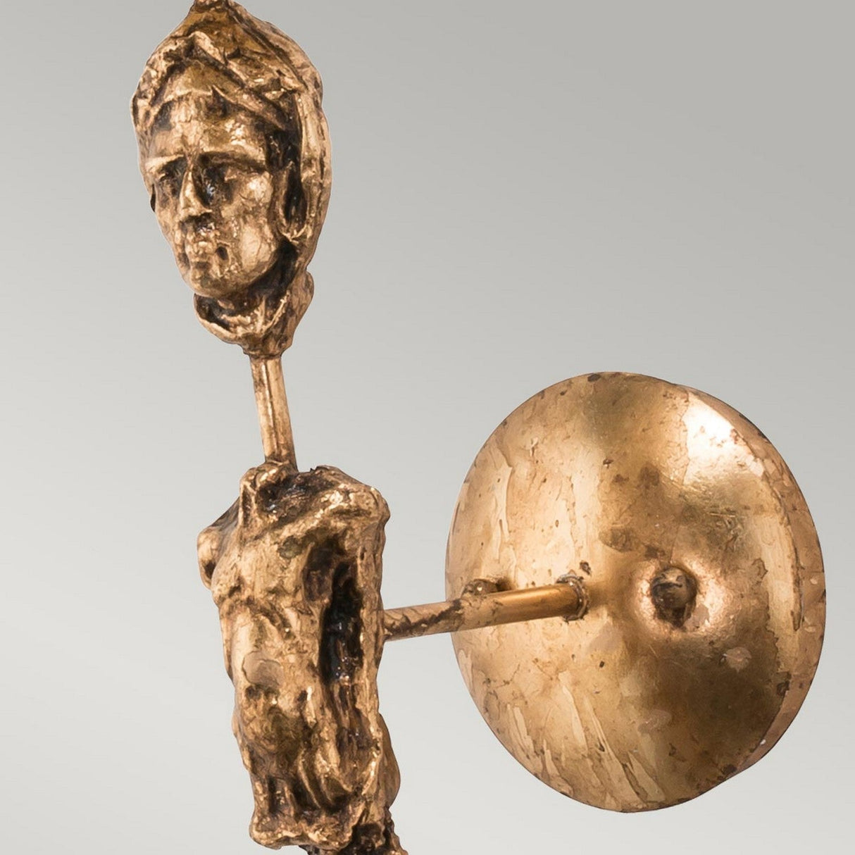 The Fragment 1 Light Wall Light - Aged Gold features a bronze sculpture with a stylized human figure, complete with an elongated body and shield. Its intricately detailed face blends modernity with vintage charm, while the abstract form boasts an aged gold finish that contrasts beautifully against the plain background.