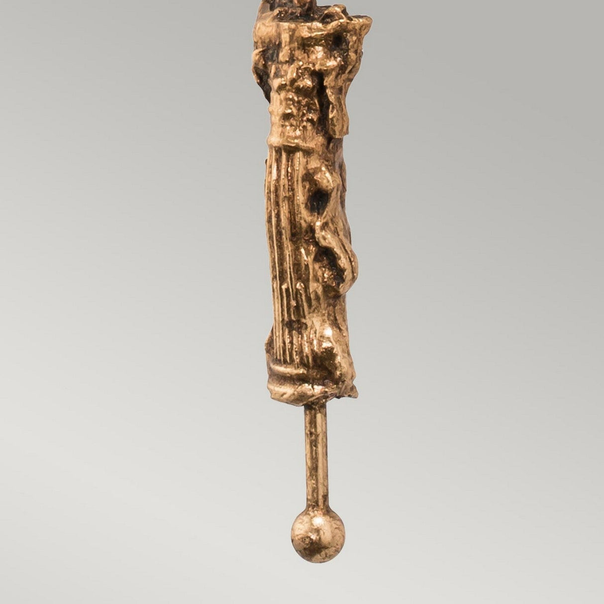 The Fragment 1 Light Wall Light in an aged gold finish features a bronze sculpture with a vertical, elongated form adorned with textured, abstract details. Its lower section tapers into a smooth cylindrical rod that concludes with a small spherical shape, blending modern aesthetics and vintage elegance against the plain gray background.