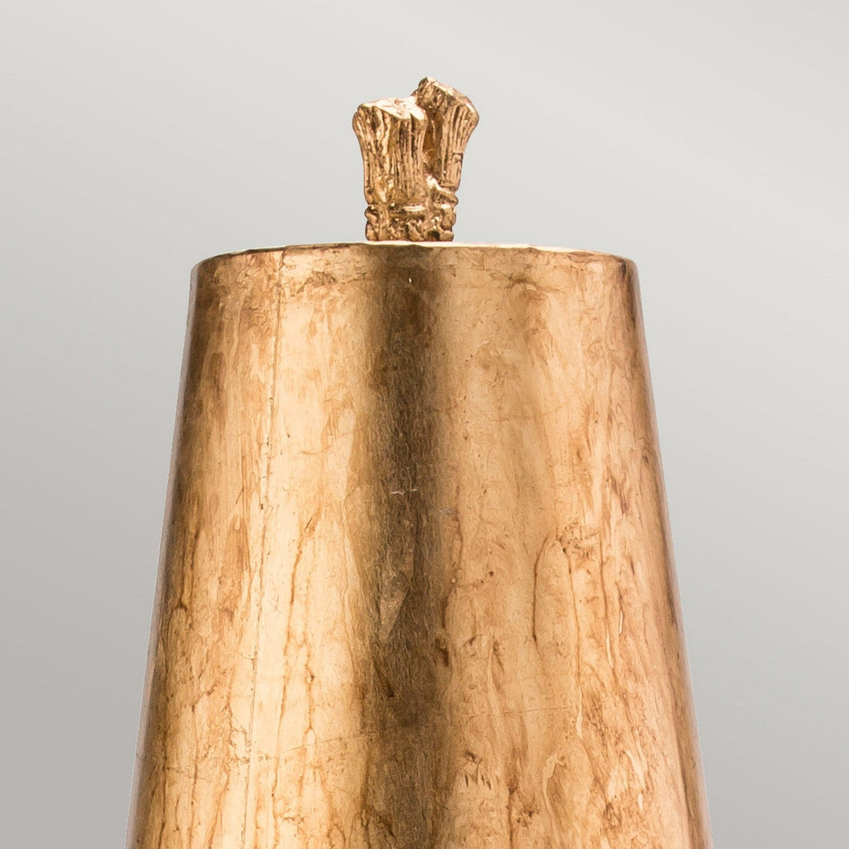 The Fragment 1 Light Table Lamp in Aged Gold boasts a tall, cylindrical design with a textured bronze surface that exudes vintage charm. Its top features an organic, branch-like shape that contrasts intriguingly with the smooth base. It stands elegantly against a neutral gray background.
