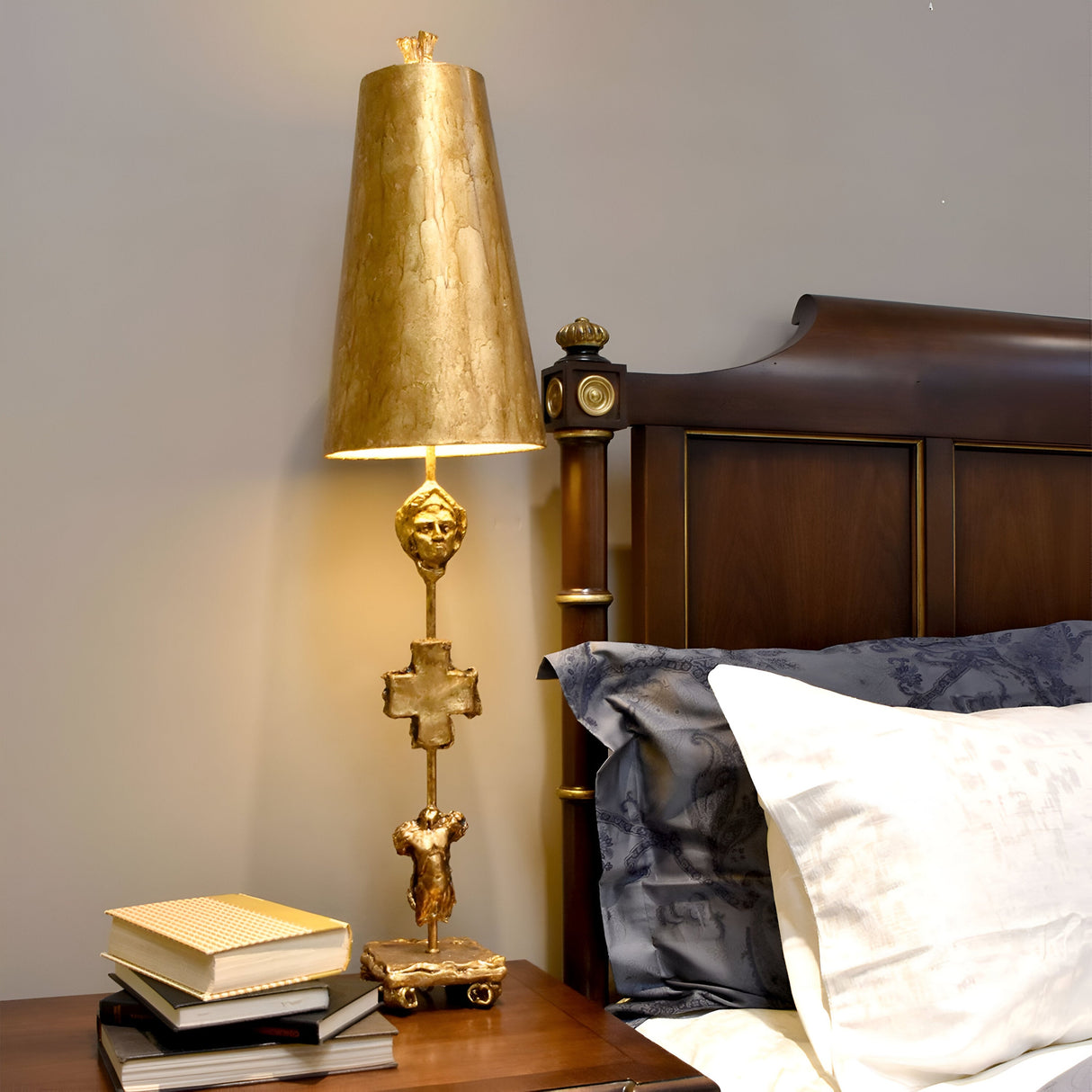 The Fragment 1 Light Table Lamp in an aged gold finish stands on a bedside table next to a wooden headboard. Its intricate design and cone-shaped shade exude vintage charm. The table is adorned with several books and a notebook, while the bed showcases visible pillows and sheets.