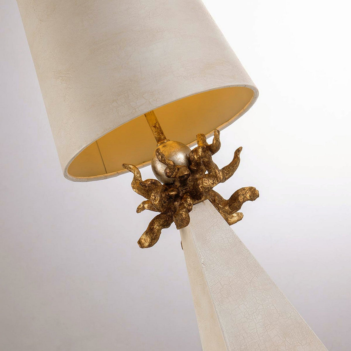 The Leda 1 Light Table Lamp features a close-up view with a tall, angled beige shade, showcasing a gold leaf design on its base that resembles flames or tendrils. This intricate design is complemented by a small metallic sphere at the center, enhancing its elegant cream patina.