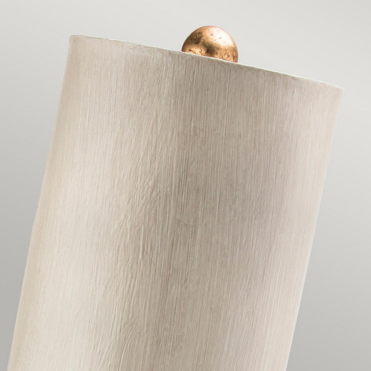 Close-up of the Madison 1 Light Table Lamp featuring a putty patina finish and an elegant, cylindrical design with a textured surface. A small, smooth sphere in gold leaf rests partially visible on top. The backdrop is a plain, muted gray.
