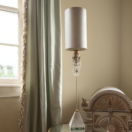 A tall, elegant Madison 1 Light Table Lamp in Putty Patina and Gold Leaf with a cylindrical shade and K9 cut glass accents stands on a glass table. It features crystal details and a geometric base. Beside it is a vintage-style wooden cabinet with ornate details. The room has light walls and draped curtains by the window.