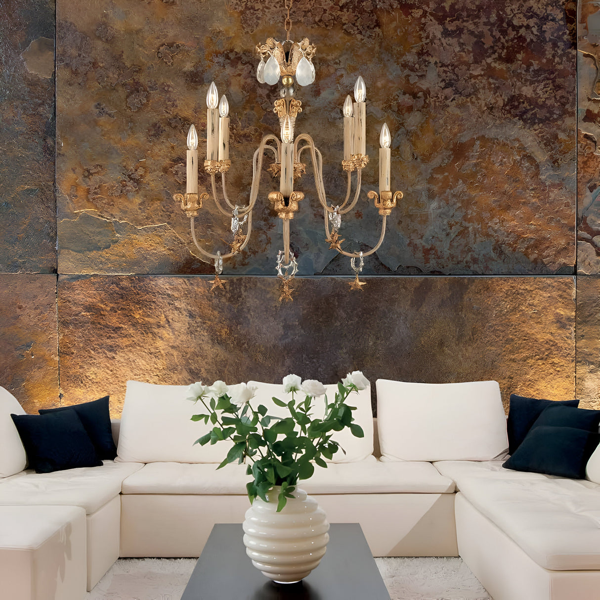Elegant living room with a white sectional sofa and black cushions. A black coffee table with a white vase holding white flowers sits in the center, illuminated by the Mignon 8 Light Chandelier - Gold & Silver adorned with crystal droplets, casting shimmering reflections against the textured, rustic wall.