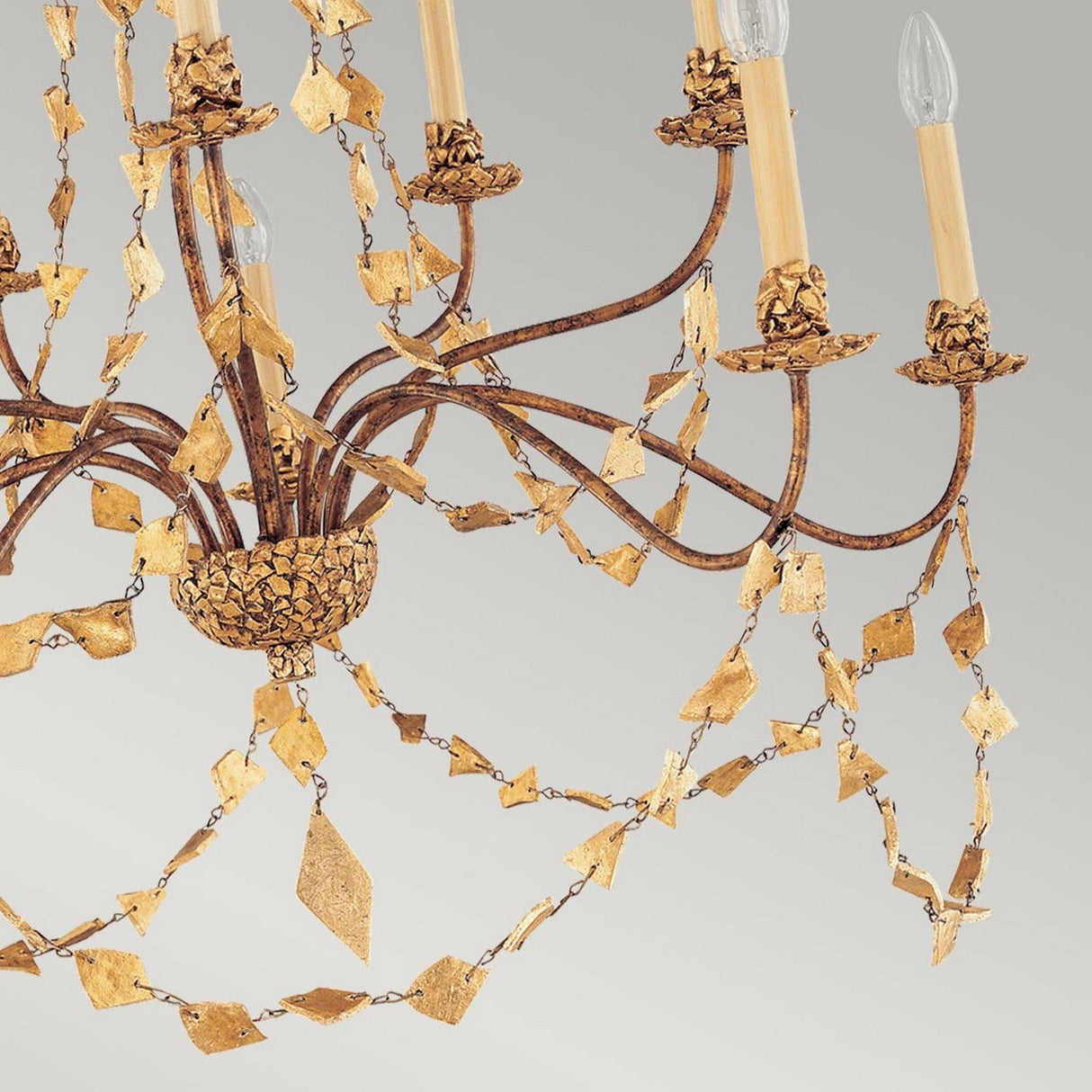 A close-up of the Mosaic 10 Light Chandelier - Gold reveals its elegant design, featuring ornate branches with leaf-like decorations and candle-style light bulbs, enhancing its vintage and luxurious appearance against a gray background.