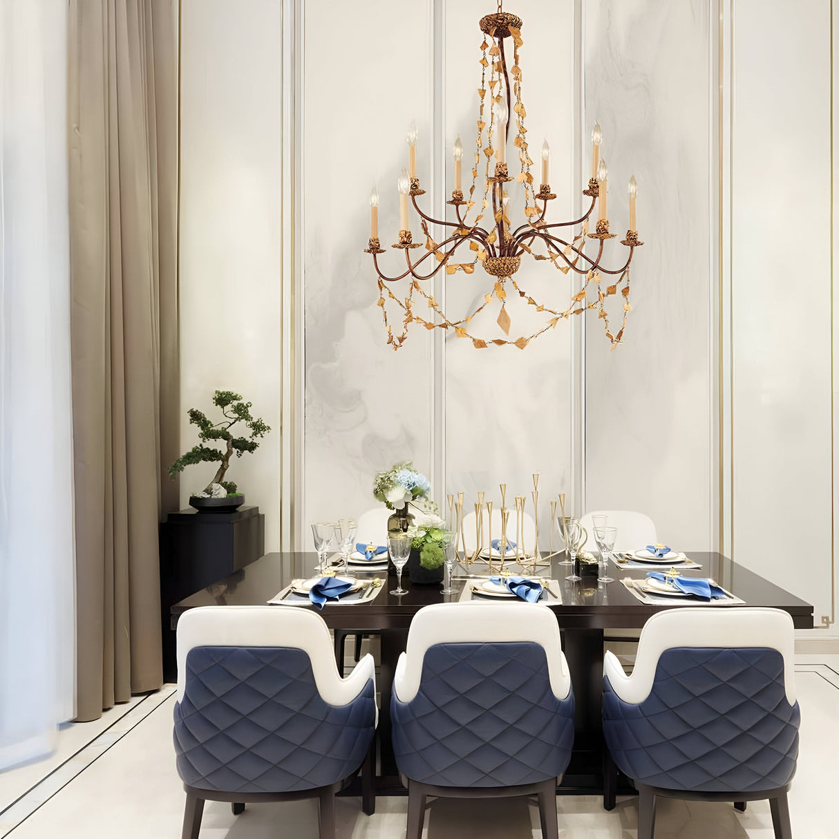 A dining room showcases a dark wooden table prepared for a meal, elegantly adorned with glassware and striking white and blue place settings. Surrounding the table are four quilted blue and white chairs. Overhead, the Mosaic 10 Light Chandelier - Gold casts a luxurious warm glow, while a small plant is placed on a nearby side table.