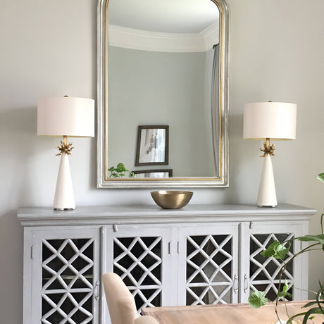 A stylish room showcases a large mirror above a gray cabinet with lattice-patterned doors, flanked by two Neo 1 Light Table Lamps in white and gold. These lamps highlight the modern design of the decorative bowl on the cabinet. A beige chair and a green plant are partially visible in the foreground.