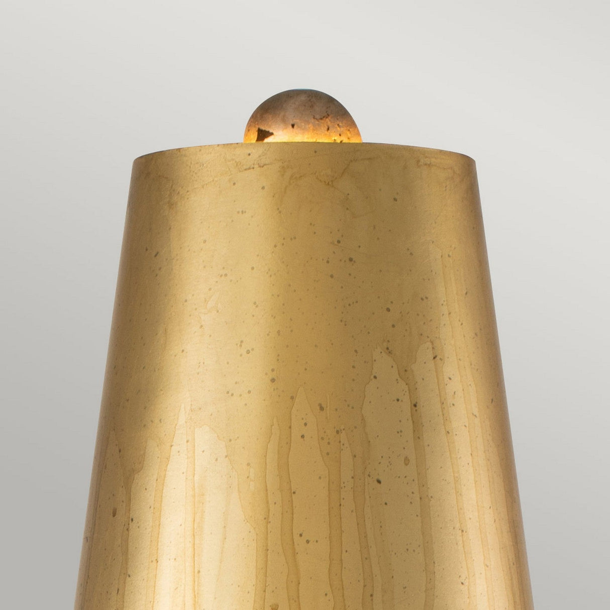 Close-up of the Noma Luxe 1 Light Table Lamp in Gold, Black & Silver Leaf, featuring a smooth, cylindrical shape with a rounded top. The surface reveals subtle vertical texturing with slight speckled patterns that reflect a soft golden hue against a plain gray background, serving as an elegant decorative accent.