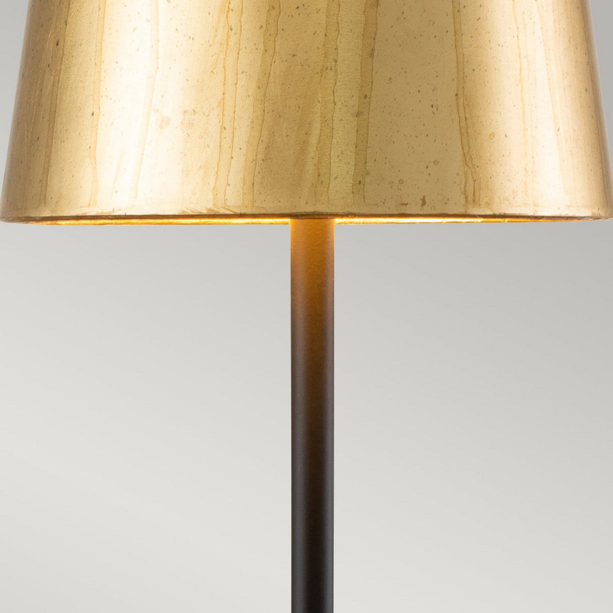 Close-up of a Noma Luxe 1 Light Table Lamp featuring a sleek black and gold stand with a beige lampshade against a light gray background. The lamp serves as a decorative accent, casting a warm glow when illuminated.