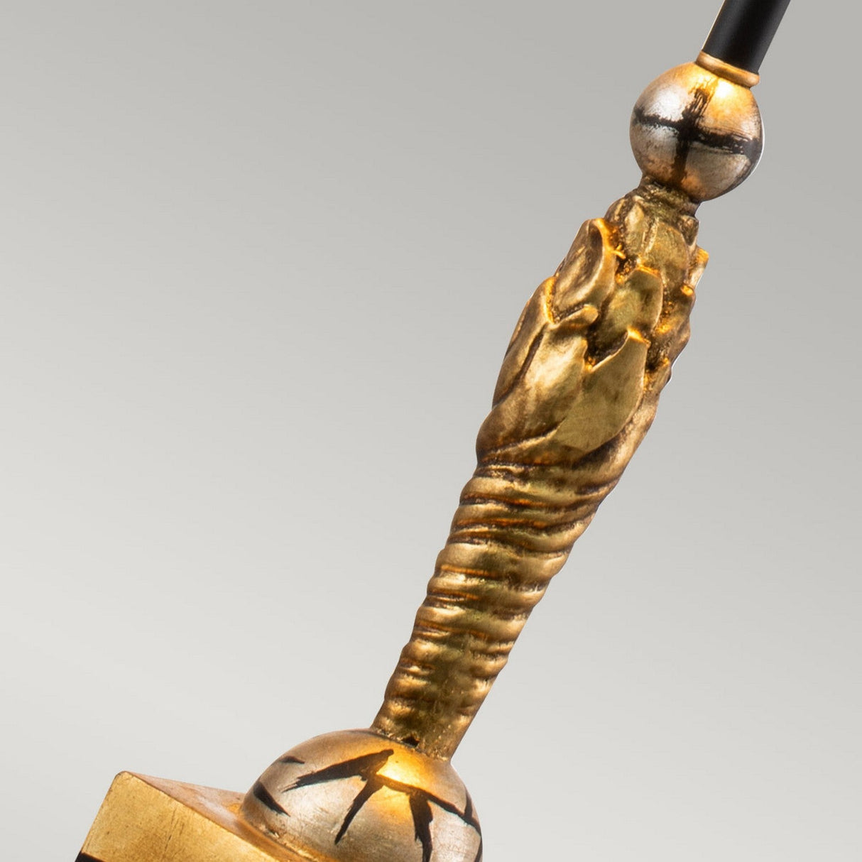 Close-up of an ornate, golden ceremonial staff with intricate detailing on the handle against a plain background. The decorative accent includes a textured grip and an elegant sphere with radial markings, reminiscent of the luxurious finish seen in the gold leaf shade of a Noma Luxe 1 Light Table Lamp - Gold, Black & Silver Leaf.