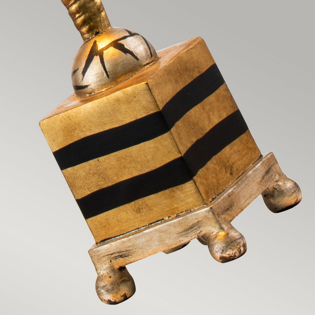 This golden block with black horizontal stripes and ornate feet, topped by a spherical accent featuring decorative scratches, serves as a decorative feature. It captures the refined elegance of the Noma Luxe 1 Light Table Lamp in gold, black, and silver leaf against the simple backdrop of neutral gray.