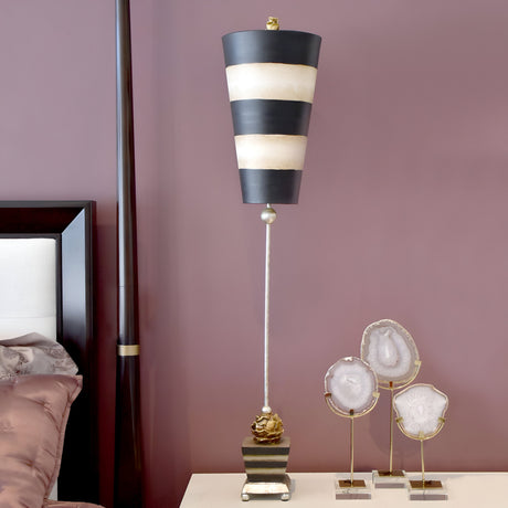 The Peony 1 Light Table Lamp with its tall, contemporary design and striped shade in taupe and black sits elegantly on a bedside table against a muted mauve wall. The lamp base showcases a stylish gold accent. Beside it, three decorative stone slices are displayed on metal stands.