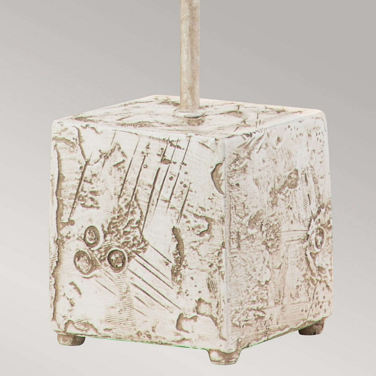 The Petra 1 Light Table Lamp - Bone White is characterized by its square stone base with abstract etching and an upward-extending metal rod. The surface boasts a weathered appearance adorned with geometric patterns, epitomizing contemporary design. Its plain, light gray backdrop accentuates the bone white base.