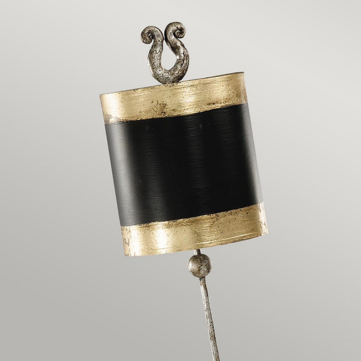 A decorative scroll-shaped ornament with a metallic base and intricate detailing, showcasing a black horizontal band and gold leaf accents, crowned with a stylized flourish that echoes the elegance of the Phoenician 1 Light Table Lamp - Black, Gold & Silver Leaf.