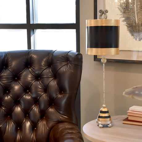 A luxurious room features a tufted dark leather armchair next to a Phoenician 1 Light Table Lamp with a black, gold, and silver leaf shade. The lamp sits on a small table adorned with glass orbs and silver leaf accents. Sunlight filters through the window, creating a warm ambiance.