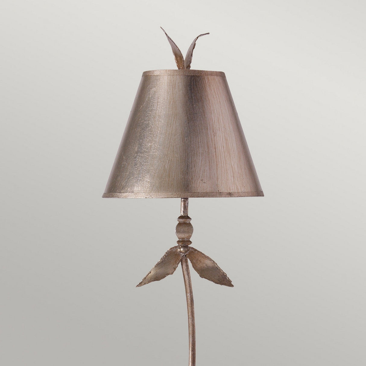 The Red Bell 1 Light Table Lamp features a decorative lamp design with a conical metallic shade in a Silver Leaf finish. It is adorned with a flower-like finial and leaf accents at the base, offering a brushed metal look that beautifully contrasts against a plain, neutral background.