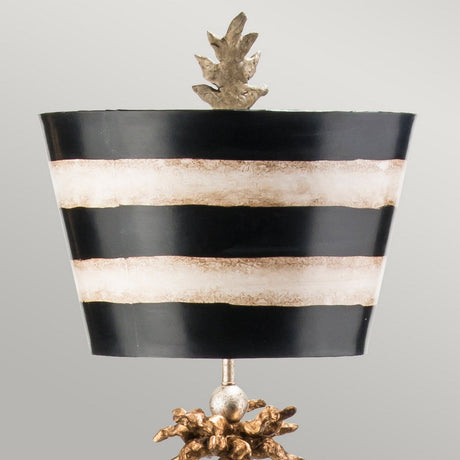 Introducing the South Beach 1 Light - Black, Putty & Gold Leaf, a piece of tropical elegance. This decorative lamp features a black and white striped lampshade with three horizontal bands, an ornate textured gold base with a star-shaped bottom, and is topped with a metallic pinecone ornament. Set against a light gray background.